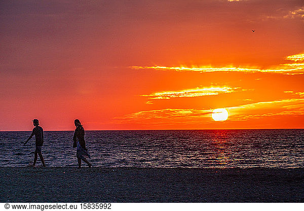 Young couple silhouette walking the beach as sun sets under a red sky.