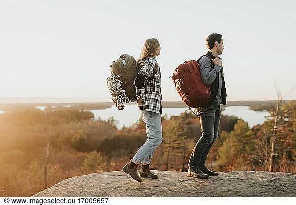 Young couple standing at edge of rocky outcrop admiring surrounding forest at autumn sunset