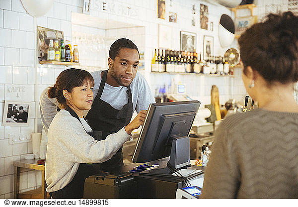 Young employee standing by female cashier using computer with customer at checkout counter in delicatessen