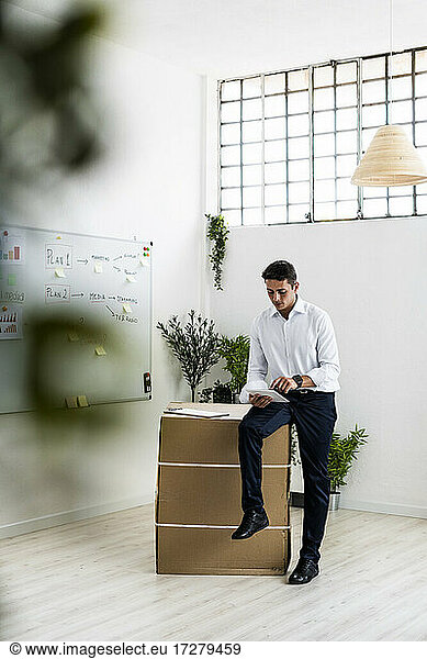 Young entrepreneur holding clipboard while sitting on cardboard box in office