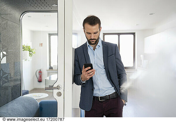 Young entrepreneur using mobile phone while leaning on telephone booth at office
