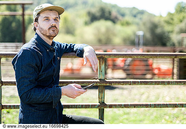 Young Farmer looking into distance holding smart phone leaning on gate