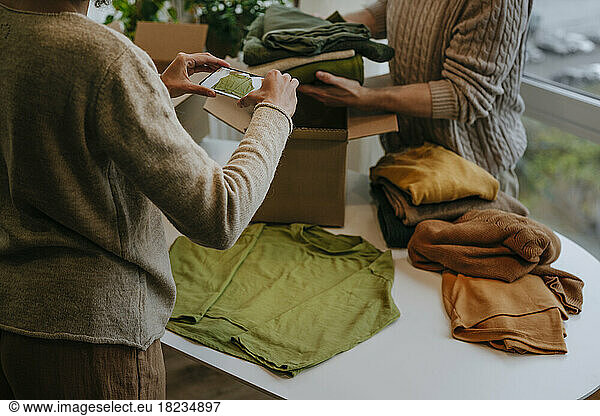 Young fashion designer photographing through smart phone with colleague packing clothes in box
