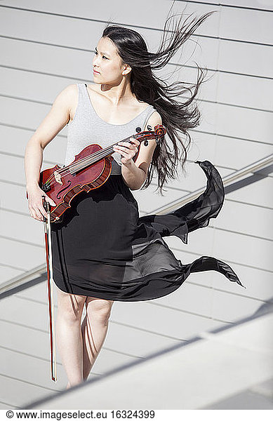 Young female Asian with blowing hair and skirt standing on stairs holding violin