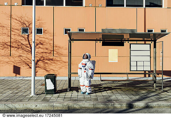 Young female astronaut in space suit waiting at bus stop during sunny day