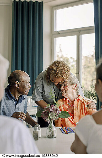 Young female caregiver embracing senior woman sitting with friends at dining table in nursing home