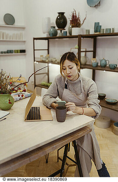 Young female entrepreneur with laptop using mobile phone while sitting at workshop