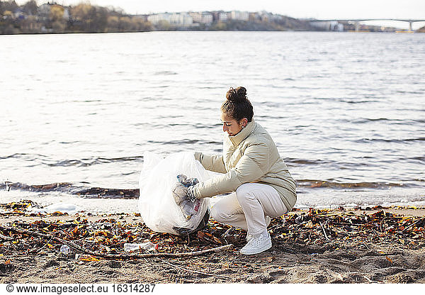 Young female environmentalist collecting waste while crouching by lake