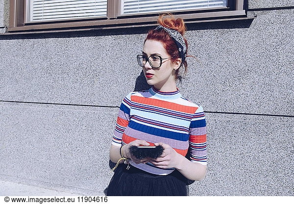 Young female hipster looking away while using mobile phone against wall