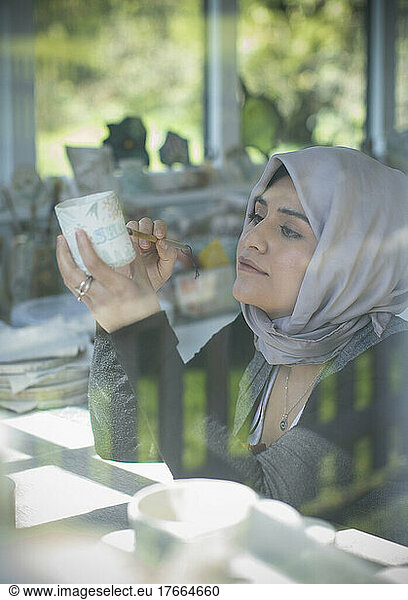 Young female Muslim artist in hijab painting ceramic mug