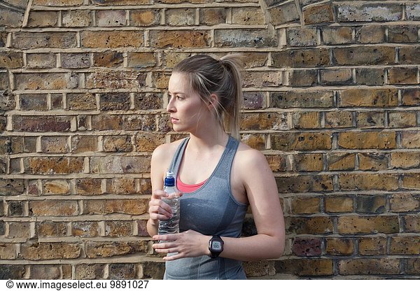 Young female runner in front of brick wall drinking water