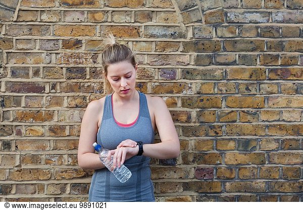 Young female runner leaning against brick wall checking stop watch