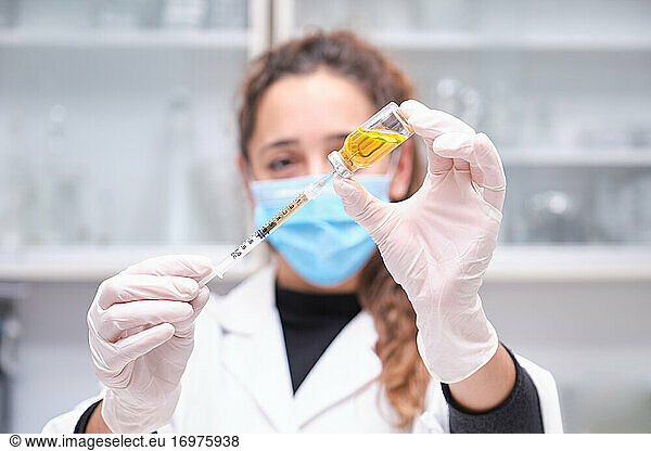 Young female scientist wearing face mask holding a coronavirus vaccine vial  syringe and needle. Covid-19 vaccine development.