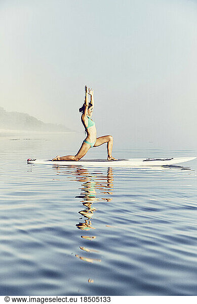 Young female standup paddle boarder doing yoga at sunrise in the fog