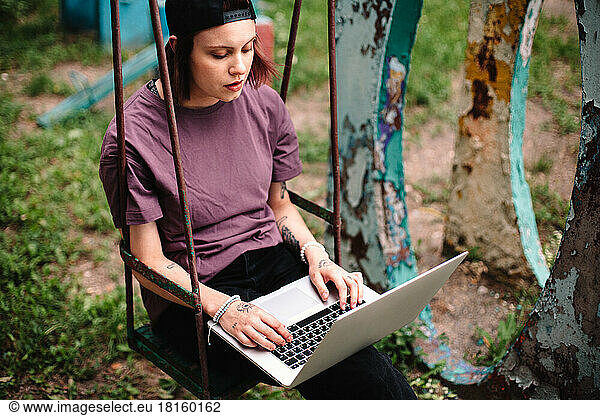 Young female student using laptop sitting on a swing in summer