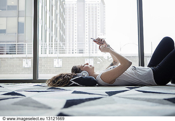 Young female using smart phone while lying on carpet by window at home