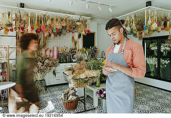Young florist writing in diary by colleague walking in flower shop