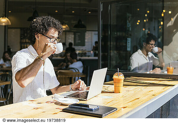 Young freelancer drinking coffee and sitting with laptop in cafe