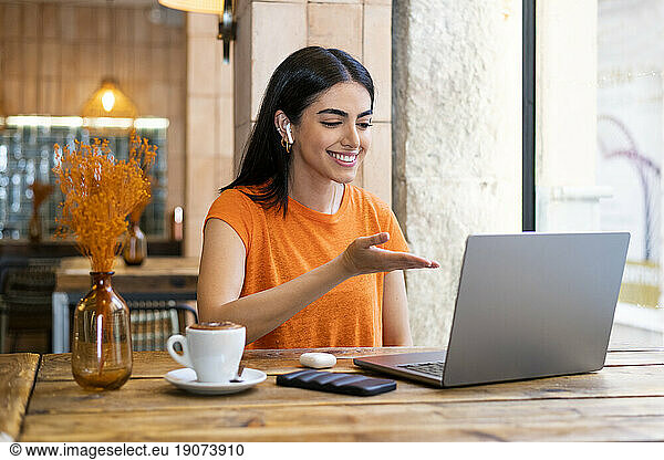 Young freelancer gesturing on video call through laptop sitting in cafe