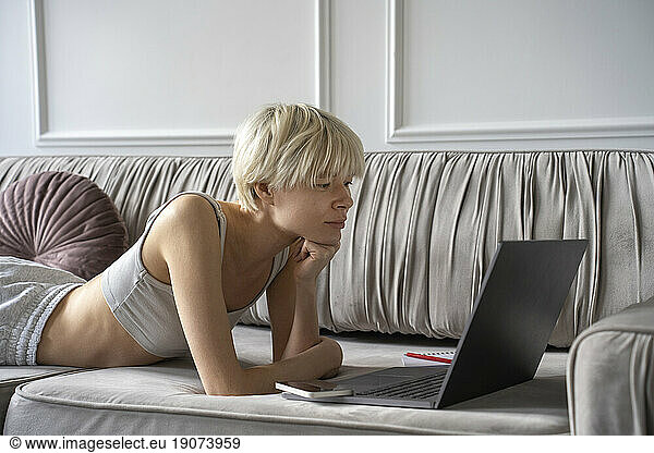 Young freelancer looking at laptop lying on sofa at home