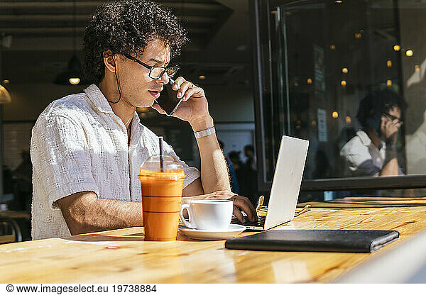 Young freelancer talking on smart phone and using laptop in cafe