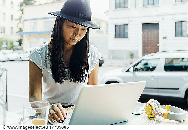 Young freelancer wearing hat using laptop sitting at sidewalk cafe