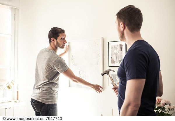 Young gay man looking at partner while hanging frame on wall at home
