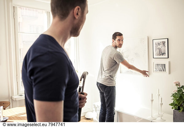 Young gay man looking over shoulder at partner while hanging frame on wall in home