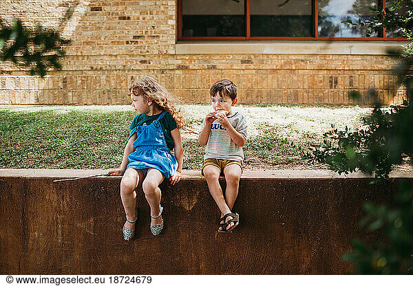 Young girl and boy sitting on wall eating together outside