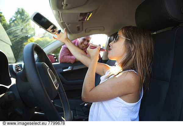 Young girl applying lip gloss pretending to be old enough to drive as her smiling father watches on on a sunny summer evening in Portland  Oregon  USA
