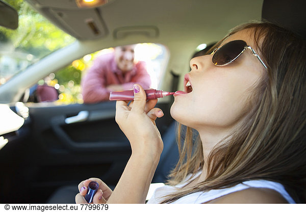 Young girl applying lip glss pretending to be old enough to drive as her smiling father watches on on a sunny summer evening in Portland  Oregon  USA
