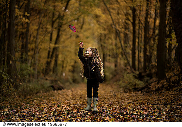 Young girl catching fall leaves in colorful forest in autumn