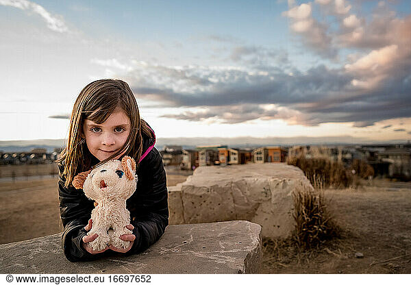 young girl holds a stuffed dog at the top of a hill at sunset