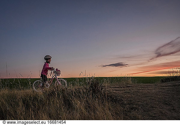 young girl on a bike waits at the top of a grassy hill at sunset
