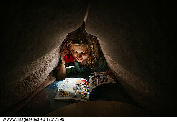 Young Girl Reading book under covers with a flashlight