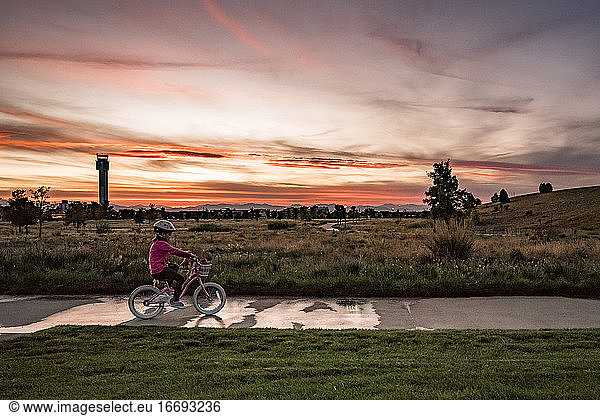 young girl rides a bike along the sidewalk in a park at sunset
