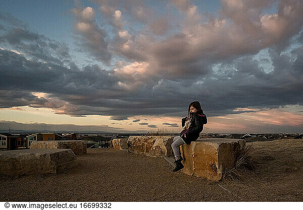 young girl sits on a rock at the top of a hill at sunset