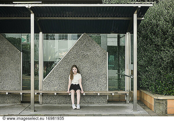 Young girl sitting alone on a wooden seat at a bus stop