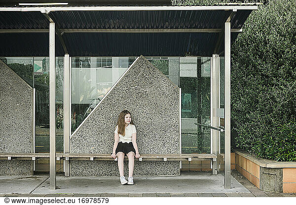 Young girl sitting alone on a wooden seat at an empty bus stop