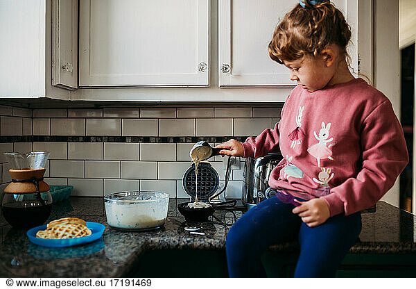 Young girl sitting on counter in modern kitchen pouring waffle batter