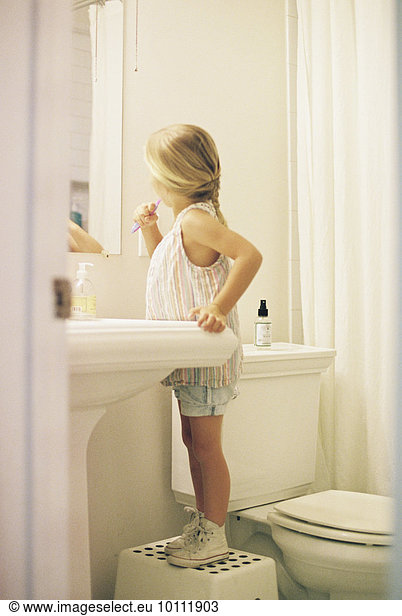 Young girl standing on a stool in a bathroom  brushing her teeth.