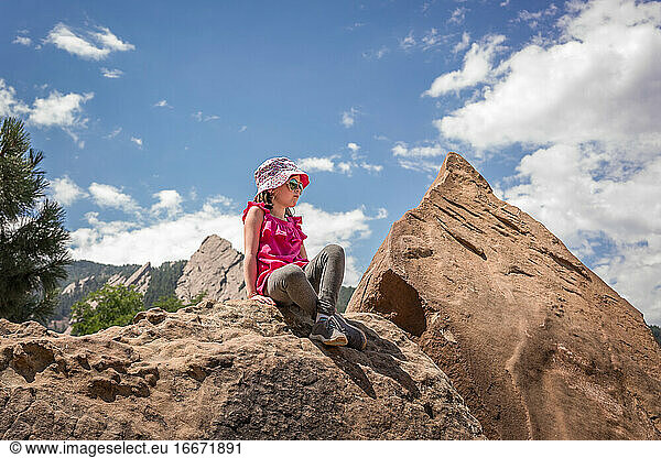 young girl with hat sits on rock with mountains behind after hiking