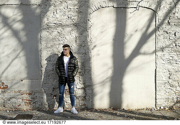 Young guy hands on pocket standing against white wall with shadows