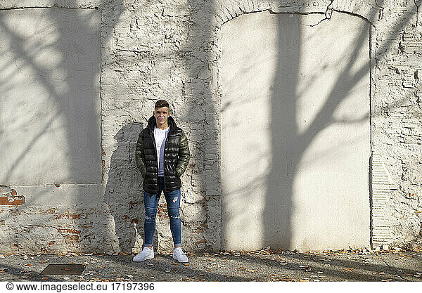 Young guy hands on pocket standing against white wall with shadows