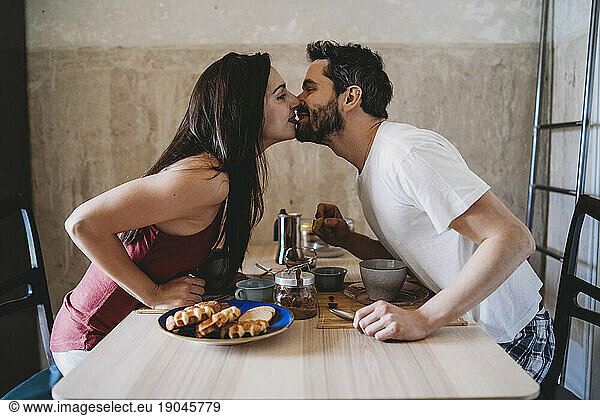 Young happy couple kisses during breakfast at the kitchen table