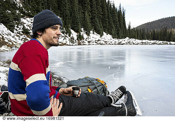 Young hockey player resting near frozen lake and drinking hot beverage
