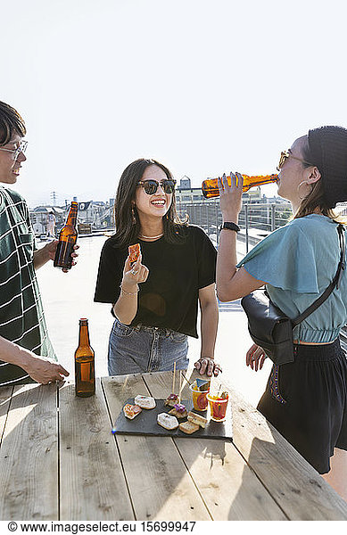 Young Japanese man and two women standing on a rooftop in an urban setting  drinking beer.