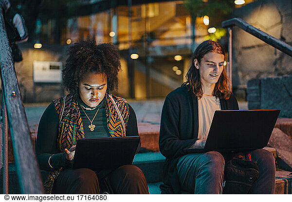 Young male and female students using laptop while sitting on steps in campus at night