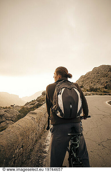 Young male backpacker with bicycle looking at mountains against sky