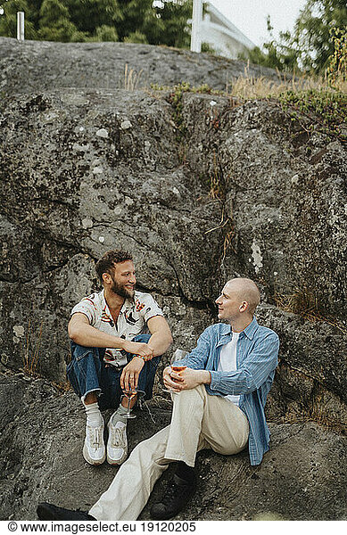 Young male friends having wine and talking to each other while sitting on rocks